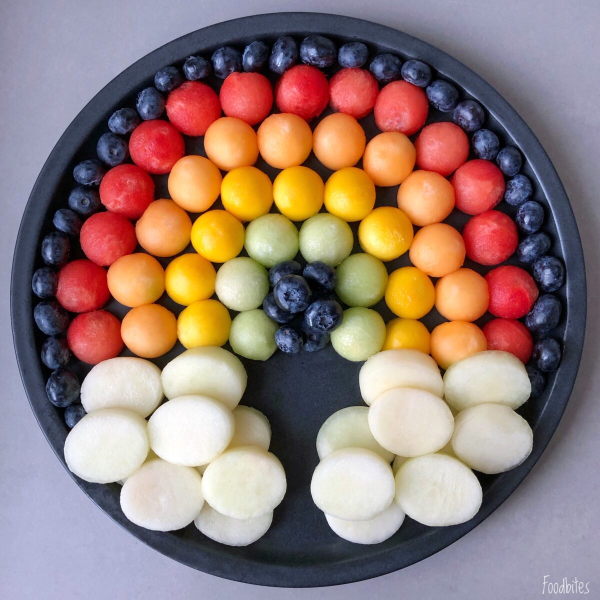 A plate arranged with melon balls in rainbow colors and blueberries, forming a rainbow shape with white melon slices as clouds at the base.