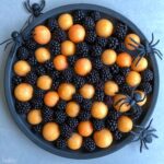 A round black tray becomes a spooky Halloween Fruit Platter, filled with blackberries and orange melon balls, and decorated with four large plastic black spiders around the edge.