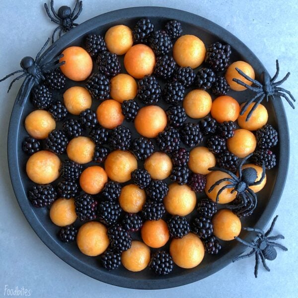 A round black tray becomes a spooky Halloween Fruit Platter, filled with blackberries and orange melon balls, and decorated with four large plastic black spiders around the edge.