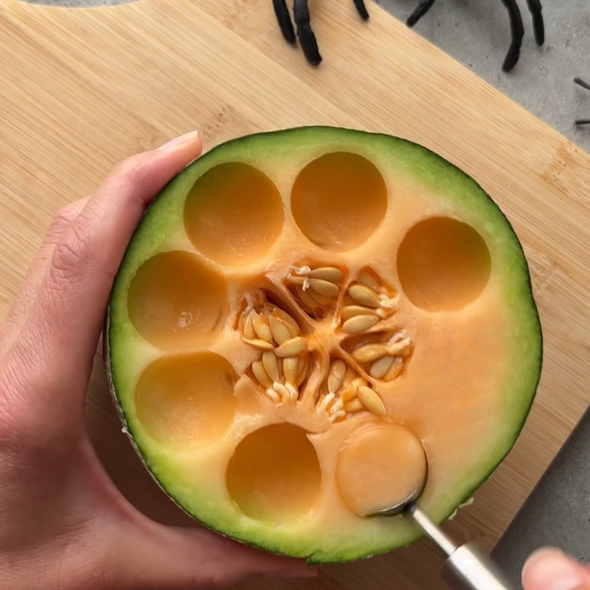 A hand uses a melon baller to scoop round pieces from a halved cantaloupe set on a wooden cutting board.