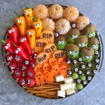 A round platter with Halloween-themed fruits and vegetables decorated with eyes, featuring peppers, carrots, kiwis, oranges, strawberries, grapes, pretzels, and cheese.