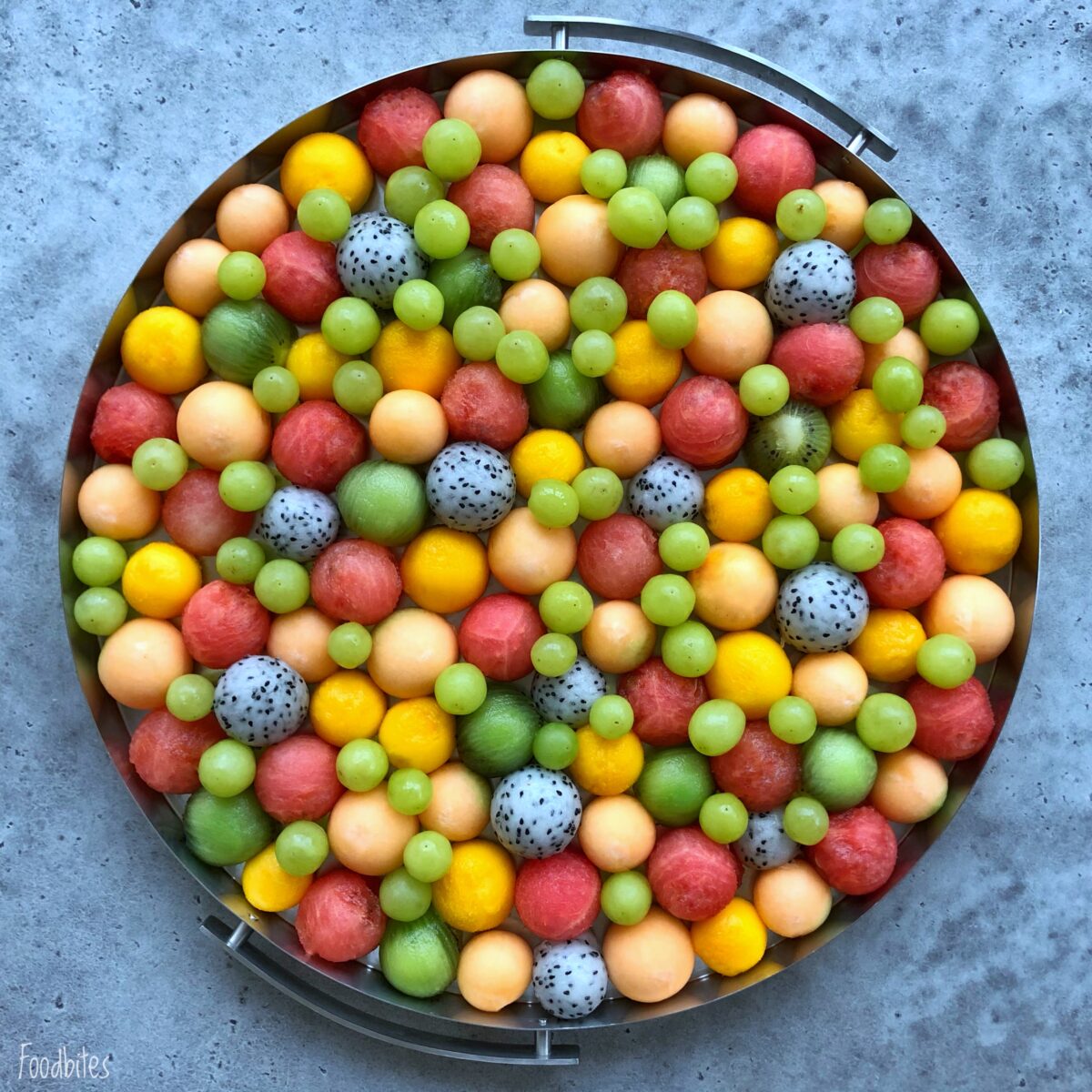 Fruit platter featuring colorful melon balls, dragon fruit balls, and green grapes arranged in a dense, even pattern on a gray surface.