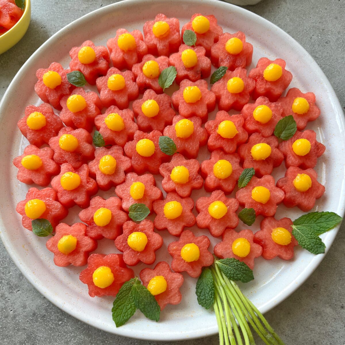 A side view of a plate of flower-shaped watermelon pieces topped with yellow mango balls, garnished with fresh mint leaves.