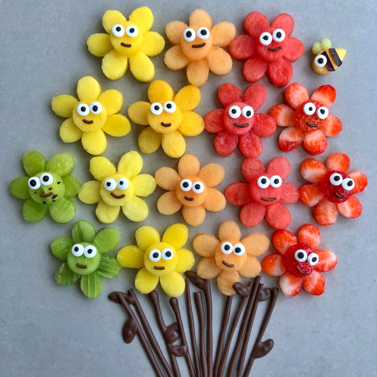 An arrangement of colorful fruit slices shaped like flowers with cartoon faces and chocolate stems. A mango bee is seen in the background.