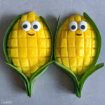 Two mango halves cut in a grid pattern, decorated with candy eyes and a smile, are arranged with green leaves to resemble smiling corn cobs.