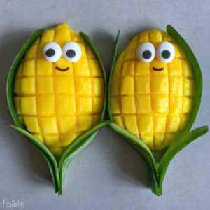 Two mango halves cut in a grid pattern, decorated with candy eyes and a smile, are arranged with green leaves to resemble smiling corn cobs.