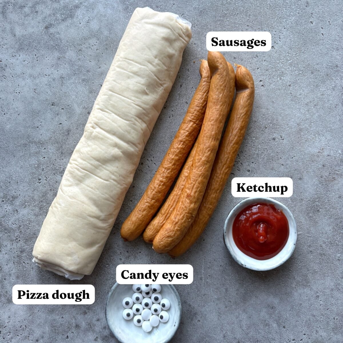 Rolled pizza dough, sausages, a small bowl of ketchup, and a dish of candy eyes arranged on a gray surface.