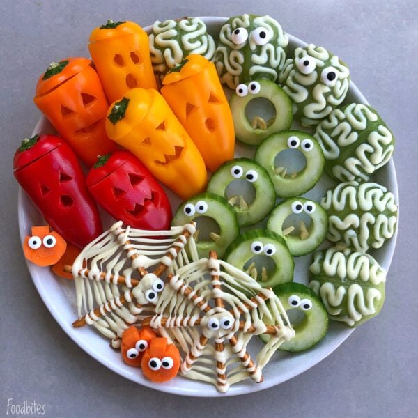 A Pretzel Spiderweb Platter features Halloween-themed veggies like carved bell pepper jack-oโ-lanterns, cucumber monsters with eyes, carrot pumpkins, and spooky pretzel and white chocolate spider webs.