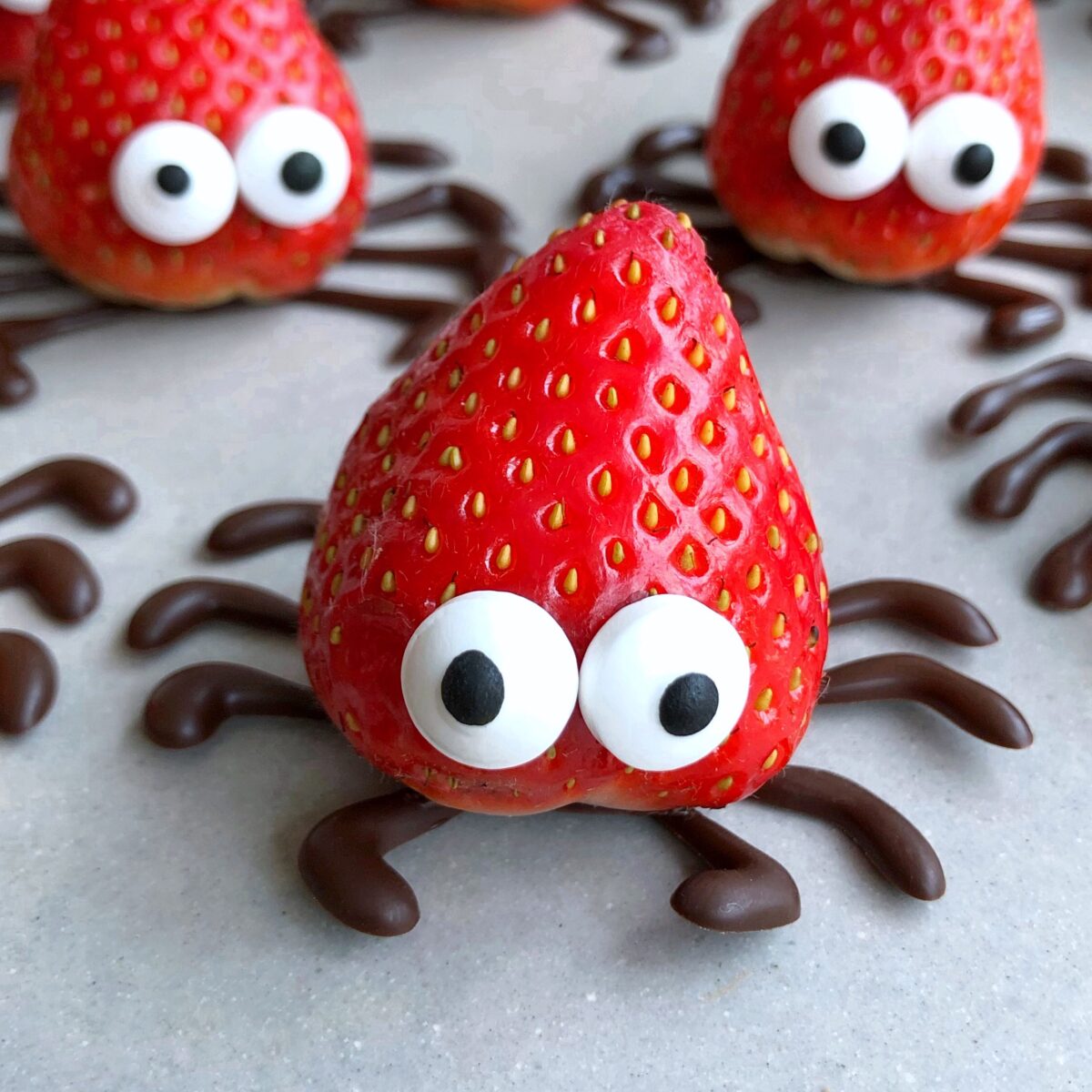 A strawberry decorated with candy eyes and chocolate legs to resemble a spider, with similar creations displayed in the background, on a light grey surface.