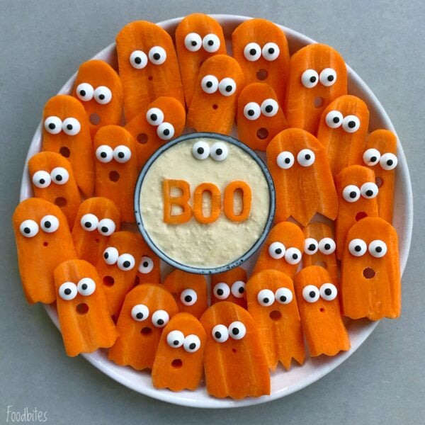 A plate of Halloween vegetables features carrot slices shaped like ghosts with candy eyes, arranged around a bowl of dip with BOO spelled on top.