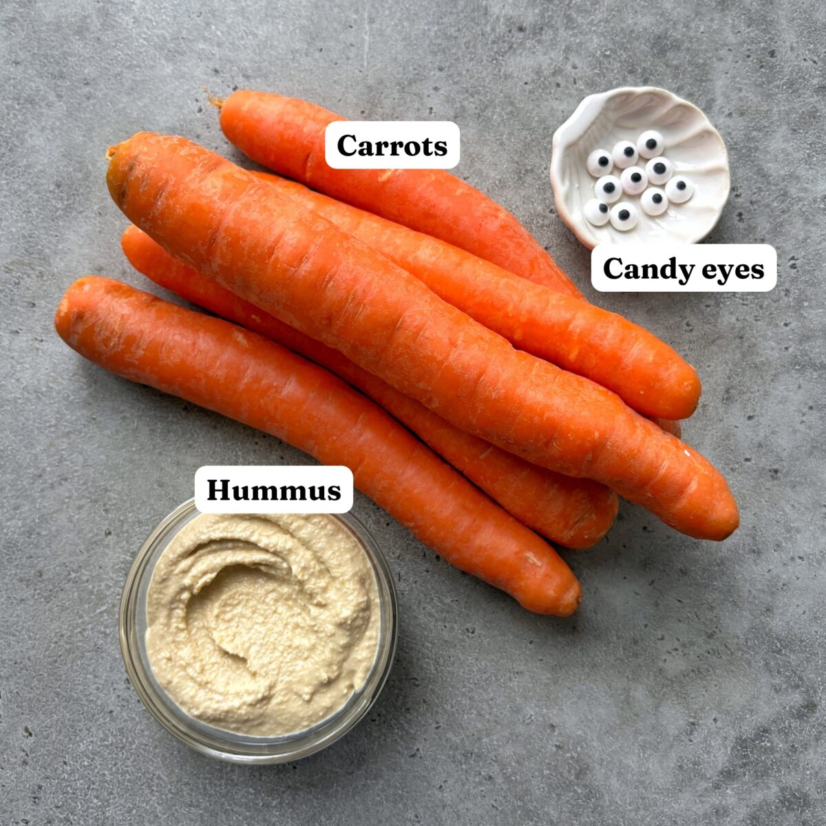 A pile of carrots, a small bowl of hummus, and a dish of candy eyes.