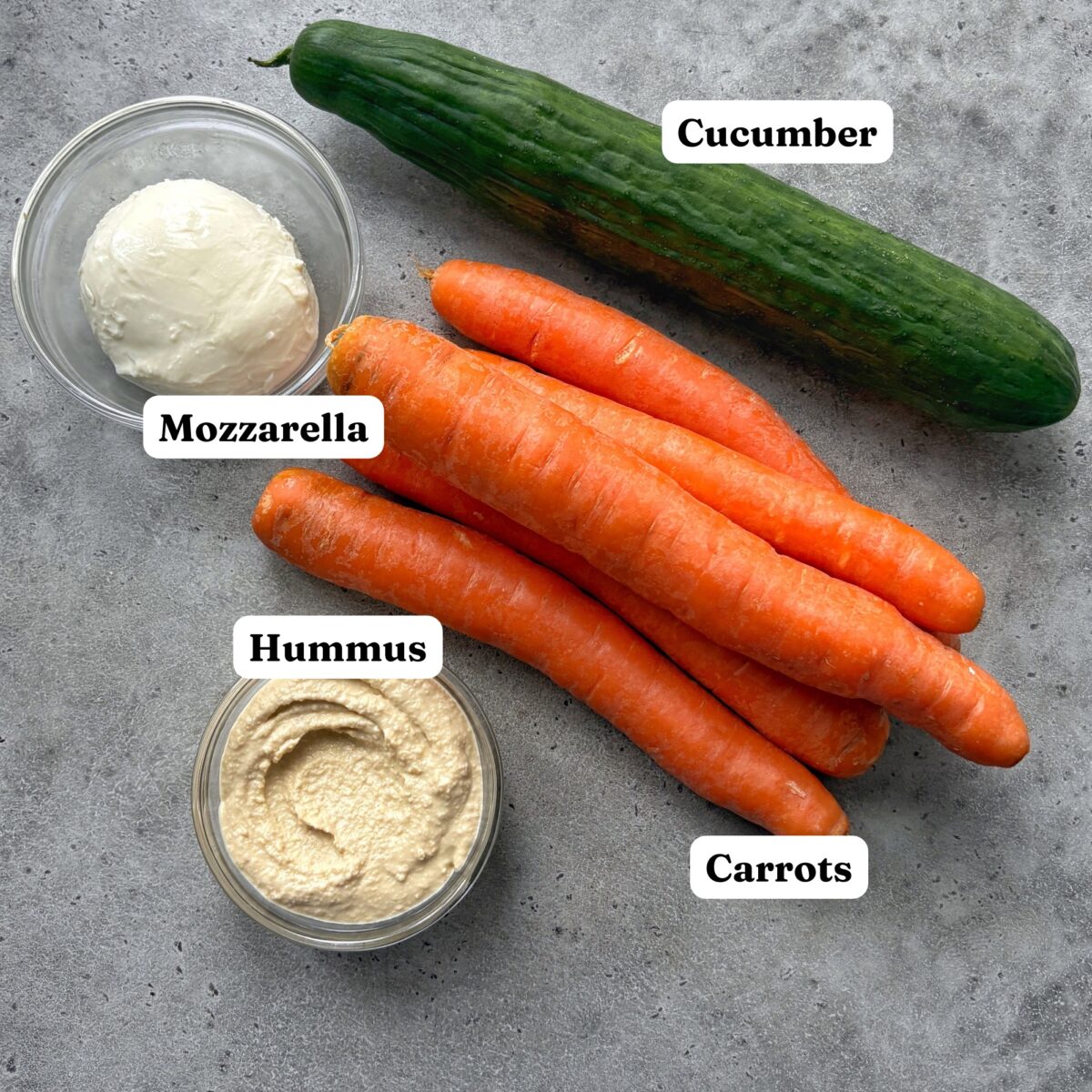 A cucumber, a cluster of carrots, a bowl of mozzarella, and a bowl of hummus.