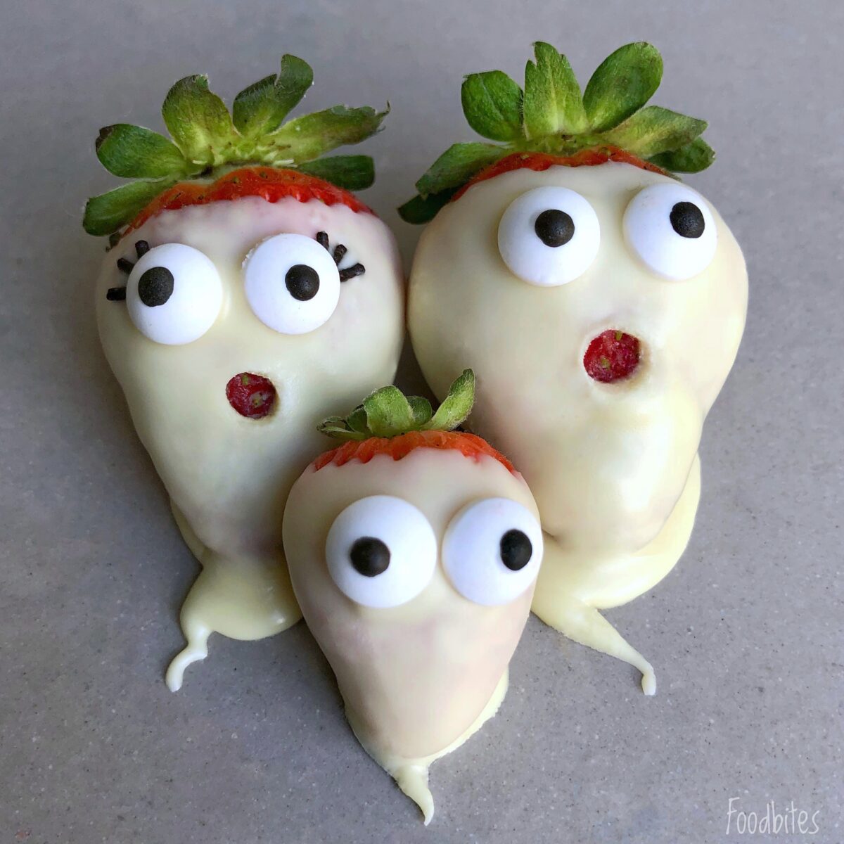 Three spooky strawberries dipped in white chocolate with candy eyes and red mouths, arranged to resemble surprised faces on a gray surface.