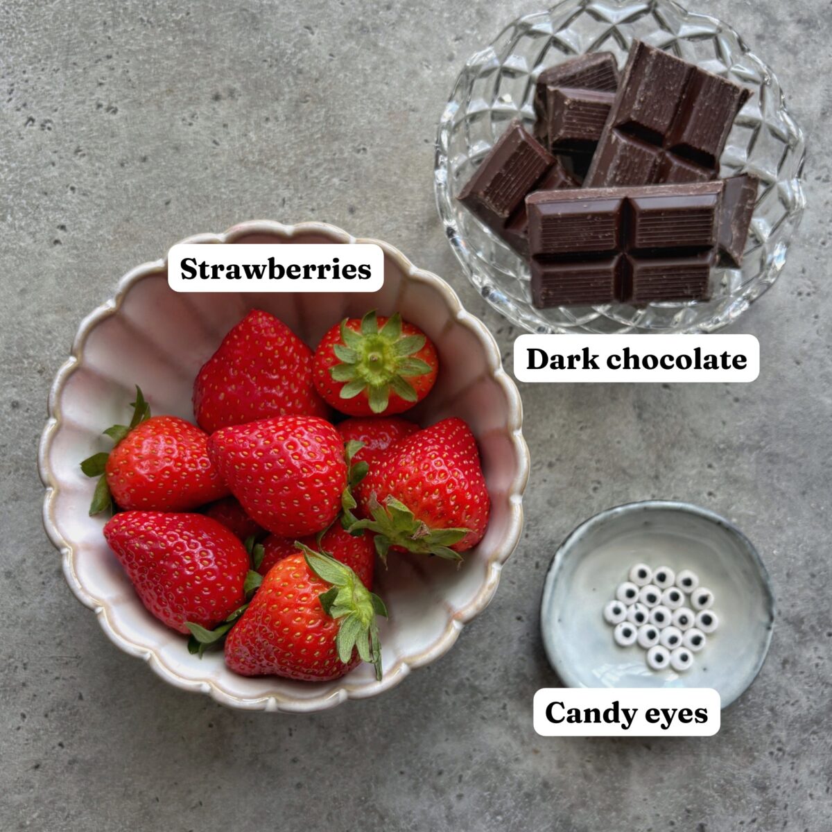 A bowl of strawberries, a bowl of dark chocolate pieces, and a small dish of candy eyes are arranged on a gray surface.