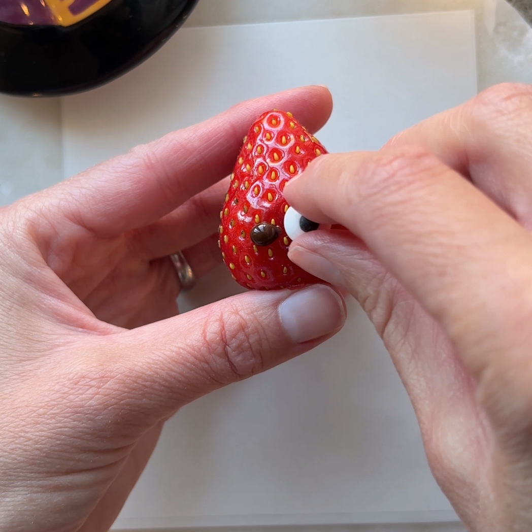 Hands stick on candy eyes to the base of a strawberry.