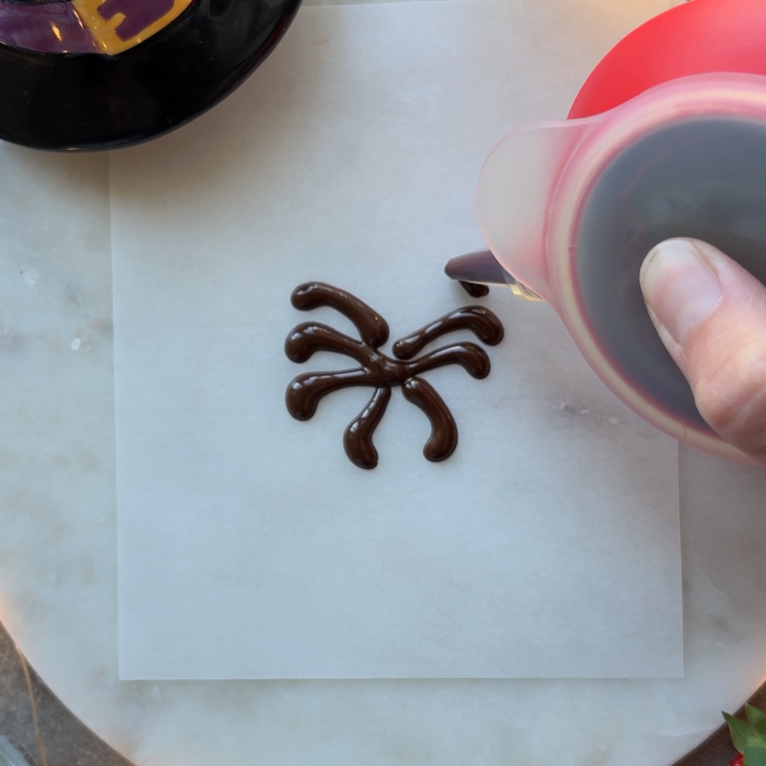 A hand pipes dark chocolate legs onto parchment paper in preparation of a Spooky Strawberry.
