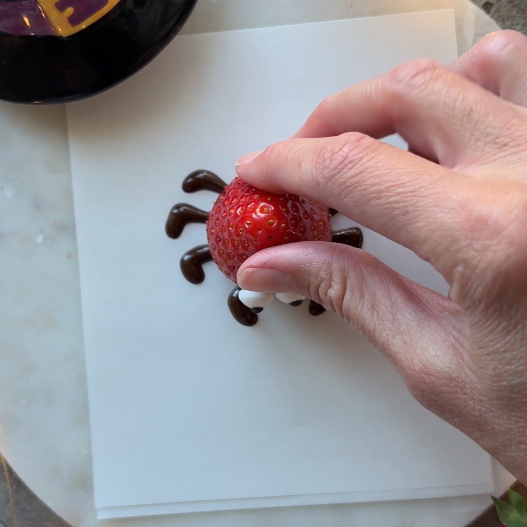 Hands top a strawberry with candy eyes onto dark chocolate legs.