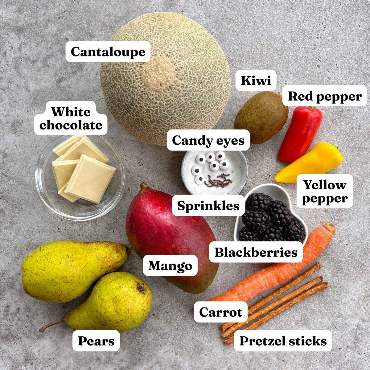 Cantaloupe, kiwi, peppers, white chocolate, candy eyes, sprinkles, blackberries, mango, pear, carrot, and pretzel sticks arranged on a grey surface.