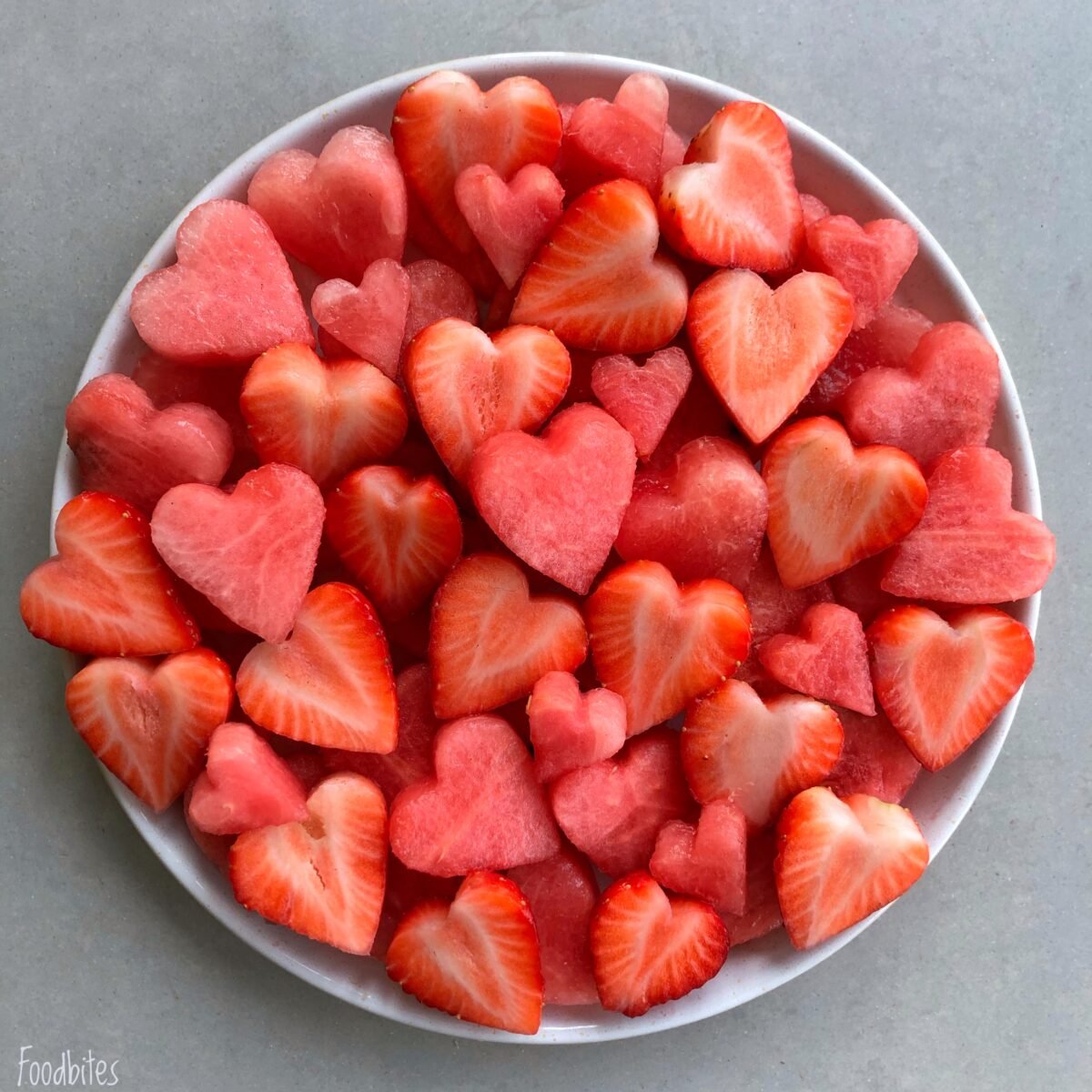 A white plate filled with heart-shaped pieces of watermelon and strawberries on a light gray surface.