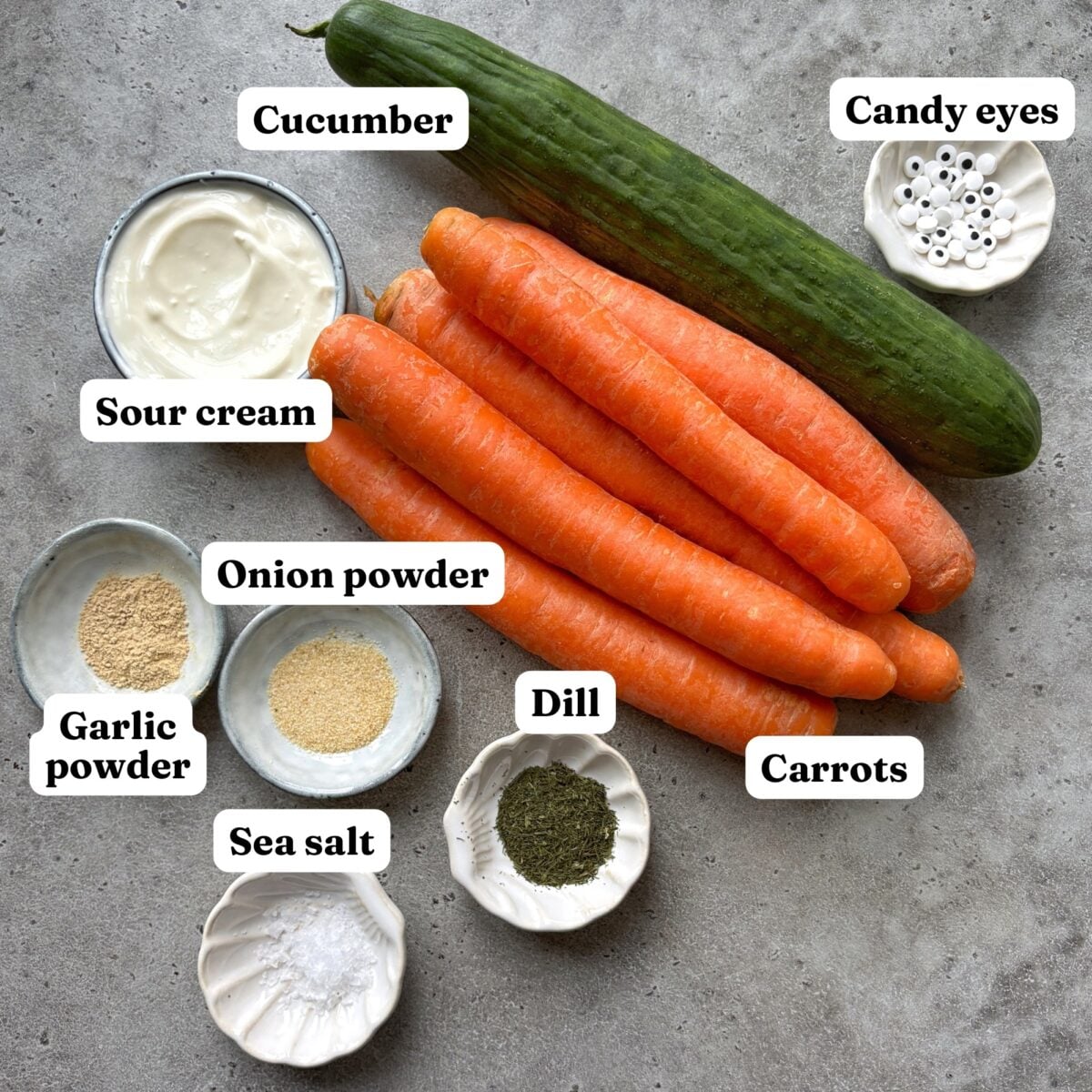 A whole cucumber, carrots, sour cream, candy eyes, and small bowls of seasoning.