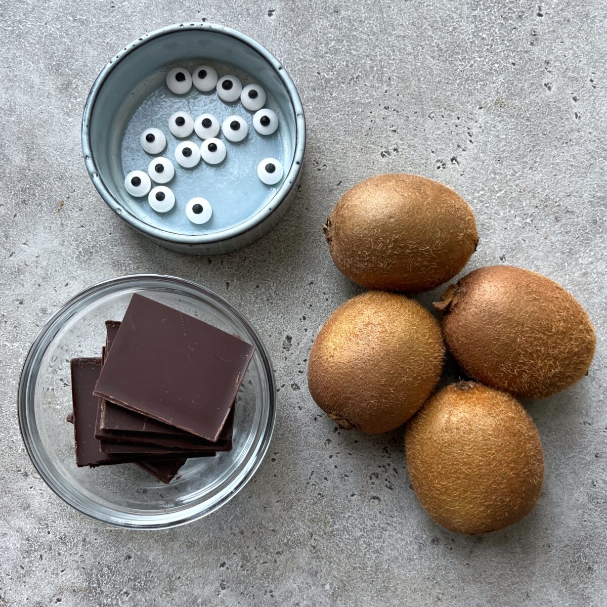 Ingredients needed for Beetlejuice Kiwis featuring a bowl of candy eyeballs, several pieces of dark chocolate in a glass dish, and four whole kiwis.