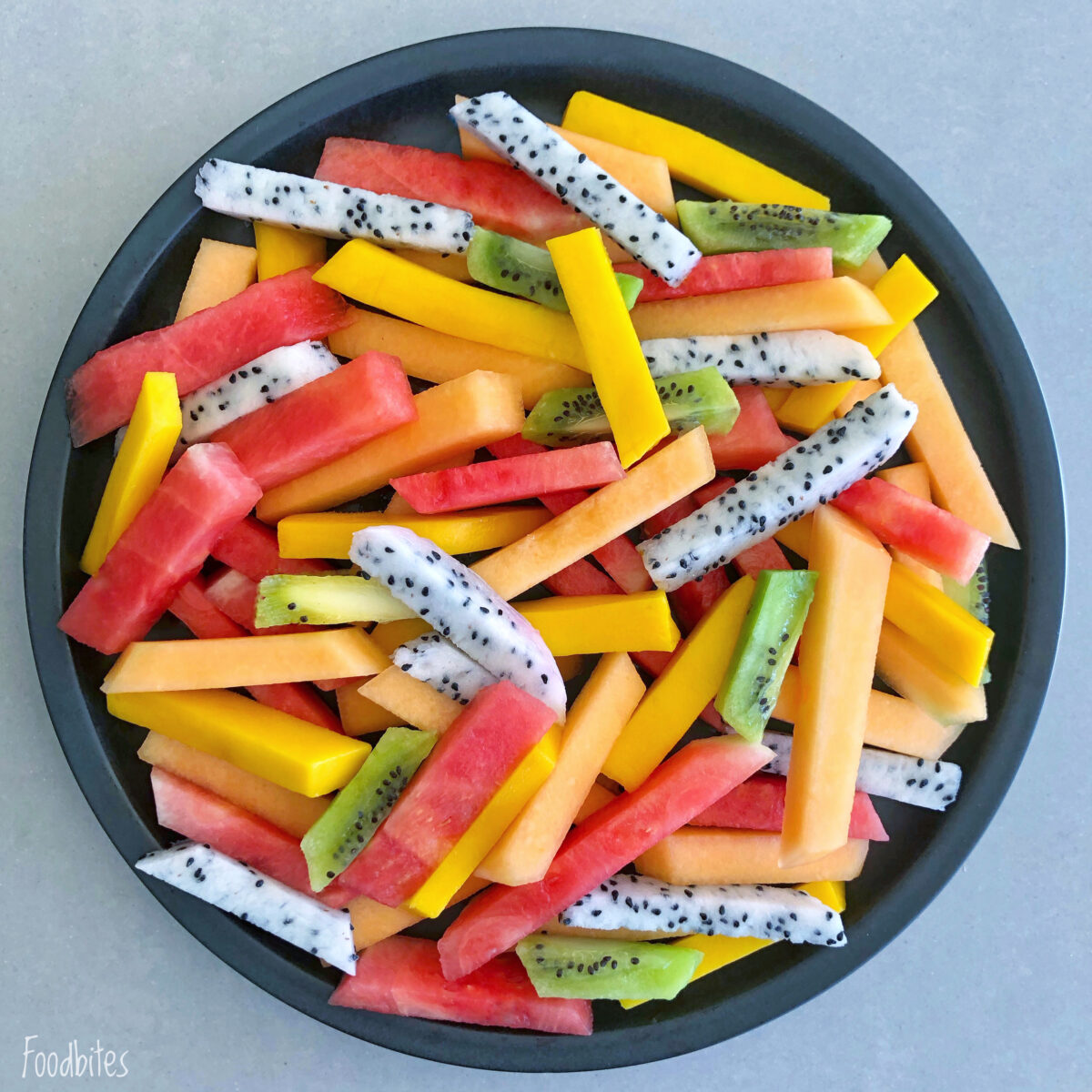 A round plate filled with neatly arranged fruit fries featuring dragon fruit, mango, cantaloupe, and kiwi sticks on a light gray surface.