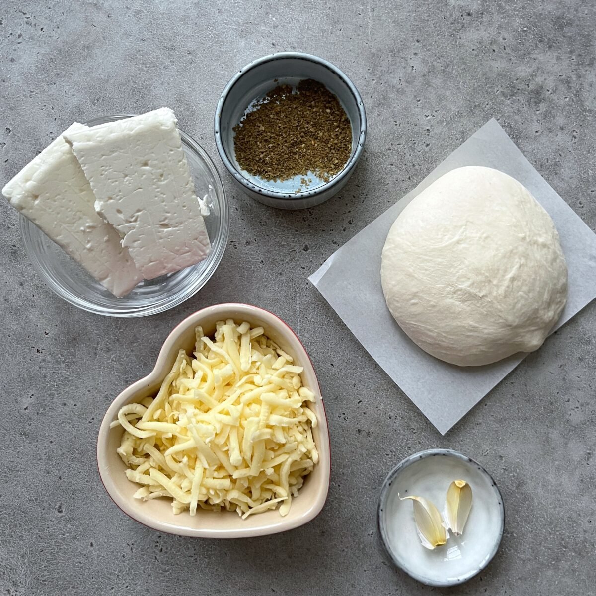 A bowl of feta cheese, a small bowl of dried herbs, a ball of pizza dough on parchment, shredded cheese in a heart-shaped bowl, and two garlic cloves on a gray surface.