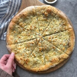 A hand holds a wooden board with a sliced Cheese Manakeesh pizza topped with herbs on a gray surface, next to a striped cloth and a small bowl of seasoning.