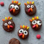 Five round chocolate protein balls decorated with colorful candy pieces to resemble cartoonish turkeys are arranged on a gray surface.
