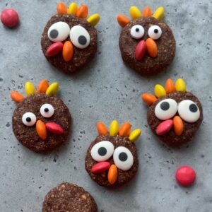 Five round chocolate protein balls decorated with colorful candy pieces to resemble cartoonish turkeys are arranged on a gray surface.