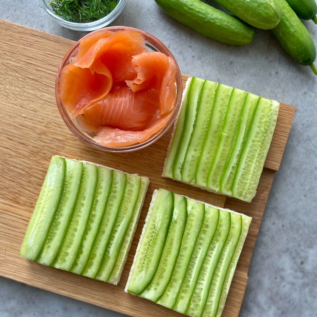 Three crustless, square pieces of bread topped with thin cucumber slices are on a wooden board, next to a bowl of smoked salmon and fresh cucumbers.