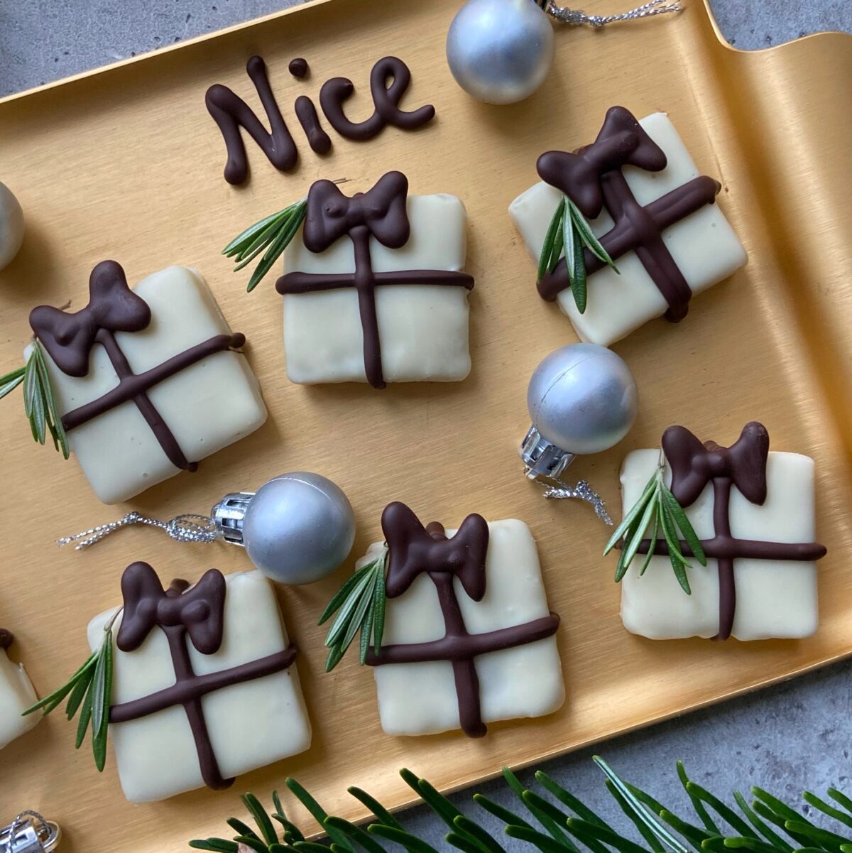 Rectangular dessert bites decorated to look like gift boxes with white chocolate coating, dark chocolate bows, and rosemary sprigs, arranged on a gold tray with silver ornaments and Nice written in chocolate.