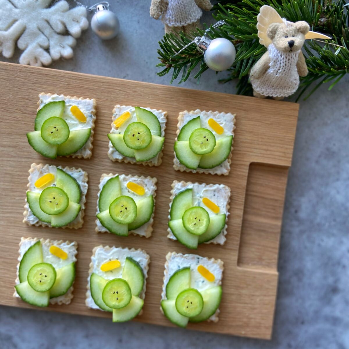 Nine square crackers topped with cream cheese, cucumber slices, and small yellow pepper halos. They are arranged on a wooden board with holiday decorations nearby.