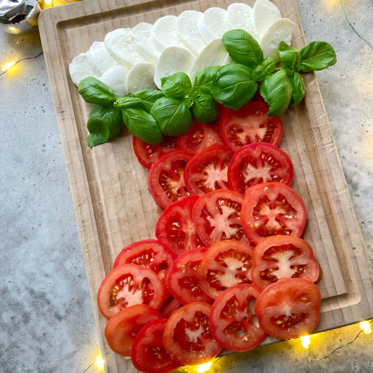 Sliced mozzarella, fresh basil, and tomato arranged in a stocking shape on a wooden cutting board, surrounded by string lights.