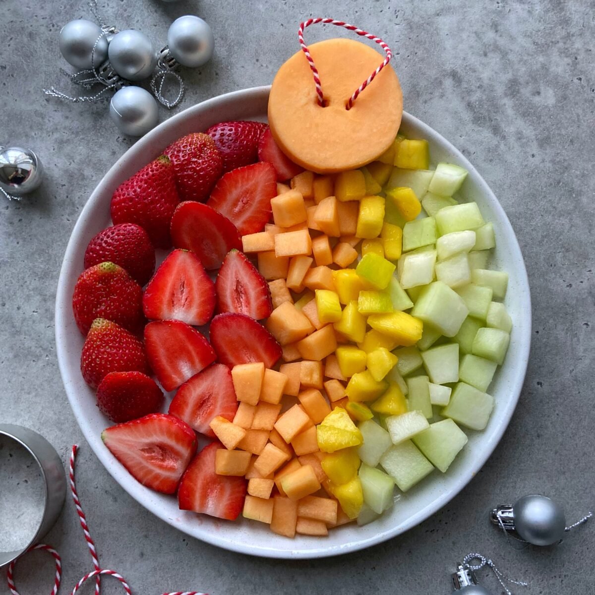 A white plate with neatly arranged sections of strawberries, cantaloupe, mango, and honeydew melon arranged to look like a bauble.