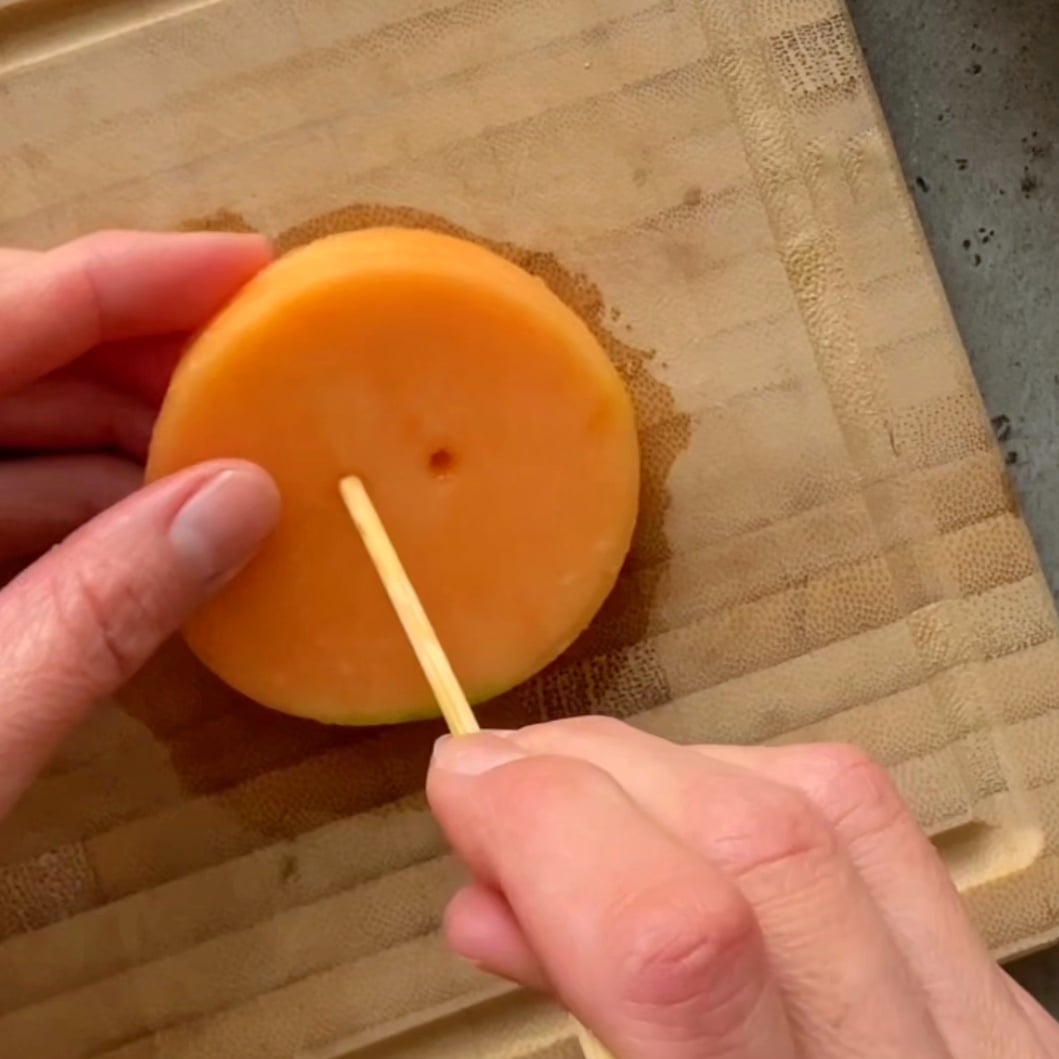 A person inserts a wooden stick into a round slice of cantaloupe on a wooden cutting board.