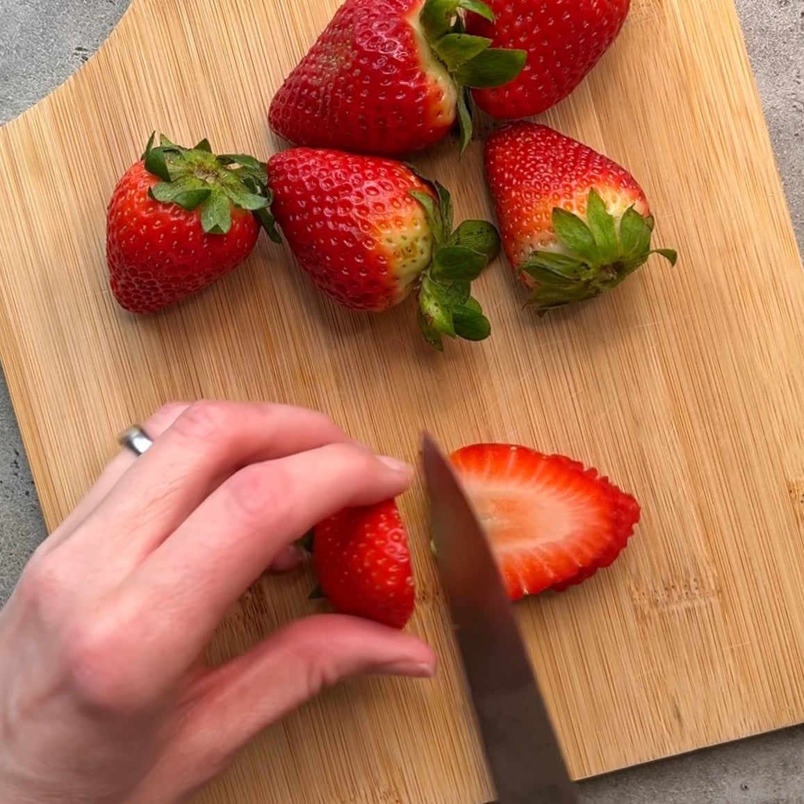 A hand slices a strawberry on a wooden cutting board with several whole strawberries nearby.
