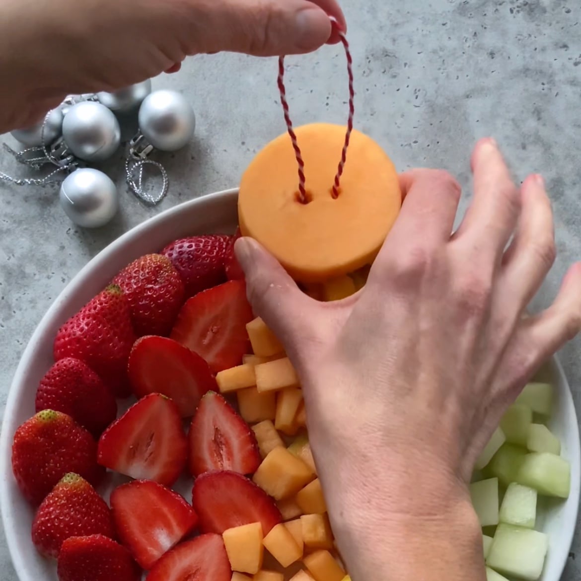 Hands arranging a Christmas Fruit Salad on a plate, with one hand holding a circular bauble made out of cantaloupe with string, next to silver ornaments on a gray surface.