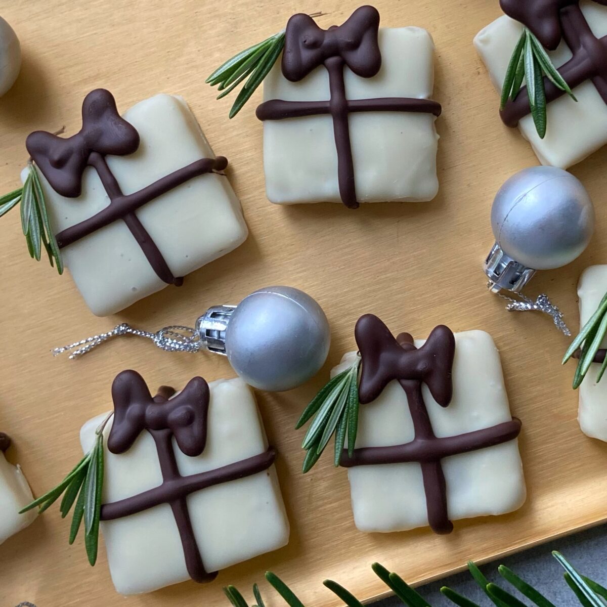 Square white chocolate Christmas treats decorated to look like gift boxes with chocolate bows and sprigs of rosemary, placed on a tray with small silver ornaments.
