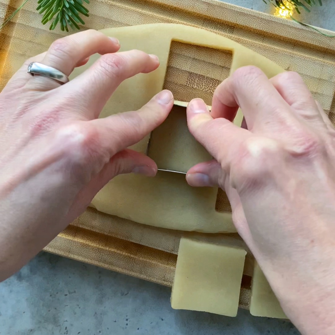 Hands using a square cookie cutter to cut marzipan on a wooden board.