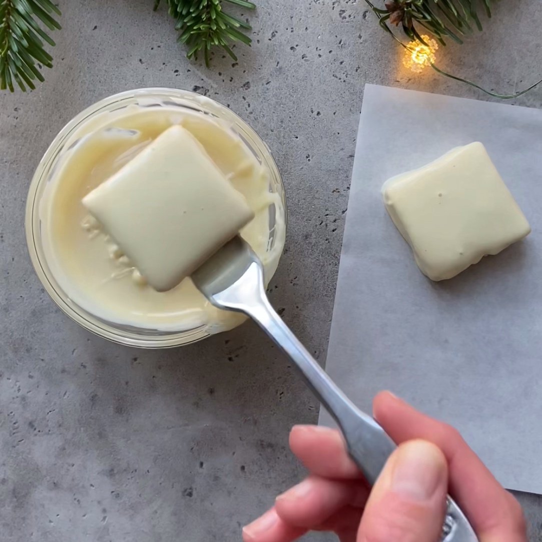 A hand dips a square Christmas treat in white chocolate using a fork, with another coated treat resting on parchment paper nearby. 