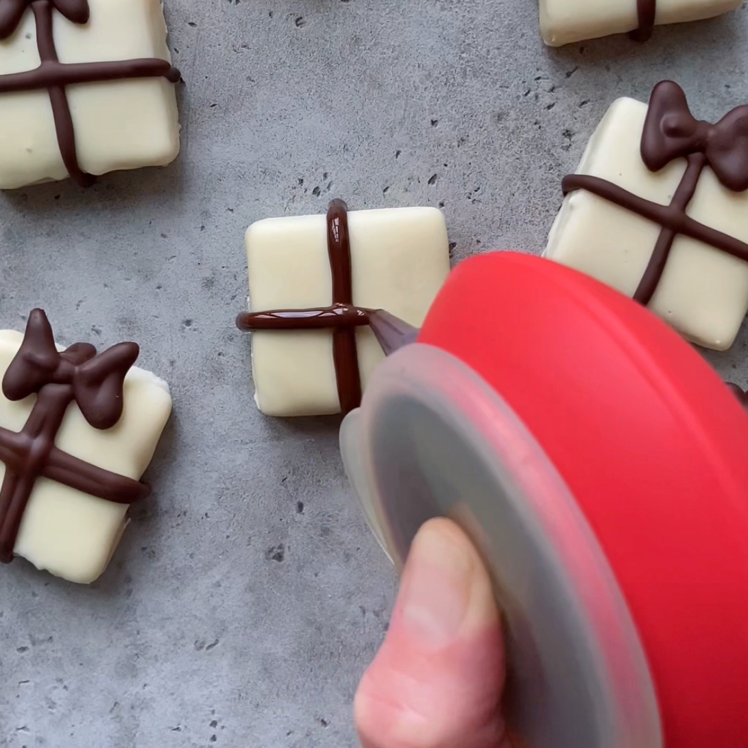 A hand decorates a white square treat with chocolate lines and a bow to resemble a gift.