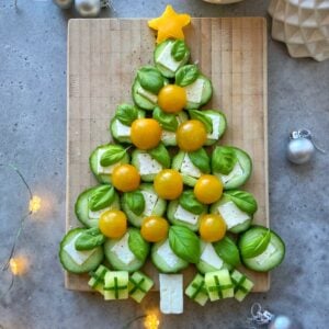Festive and healthy, this Christmas tree is crafted from cucumber slices, basil, yellow cherry tomatoes, and cheese bites on a wooden board, with little cheese and cucumber presents and a yellow star on top—perfect for holiday food recipes!.