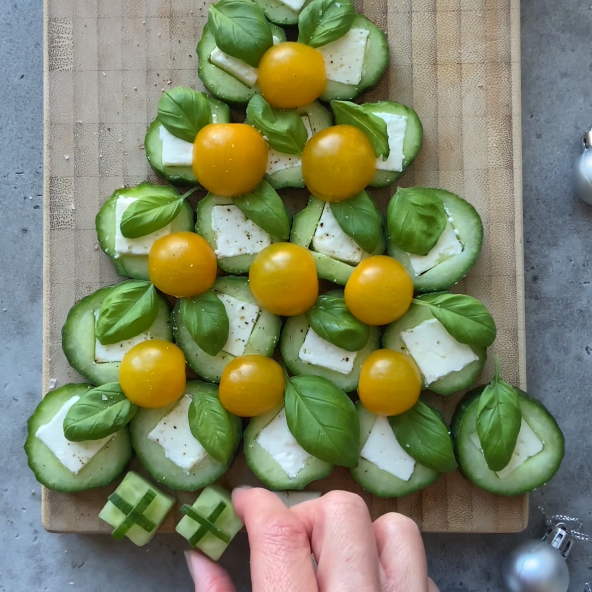 A hand places cucumber gifts underneath cucumber slices filled with feta arranged in the shape of a Christmas tree.