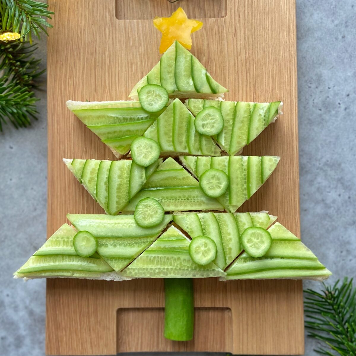 A festive bite, this sandwich is arranged in the shape of a Christmas tree, topped with cucumber slices and a yellow pepper star. Served on a wooden board.