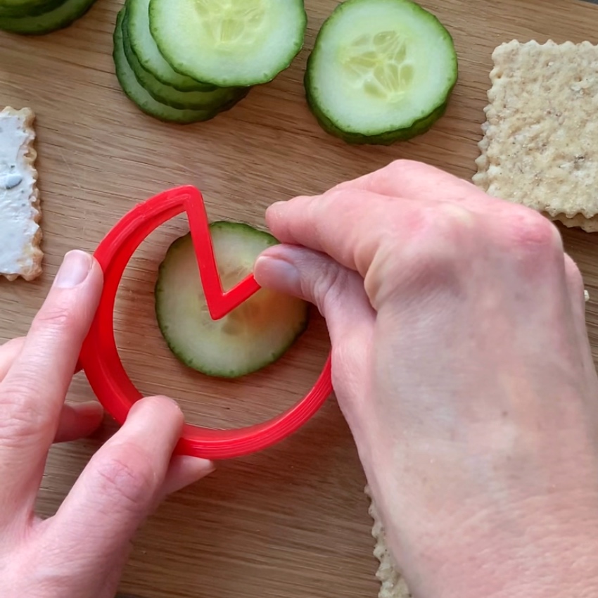A person uses a red zigzag cutter to shape a cucumber slice on a wooden board.