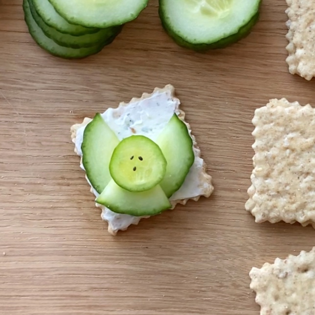 A square cracker with a layer of cream cheese, topped with cucumber slices arranged to resemble an angel.