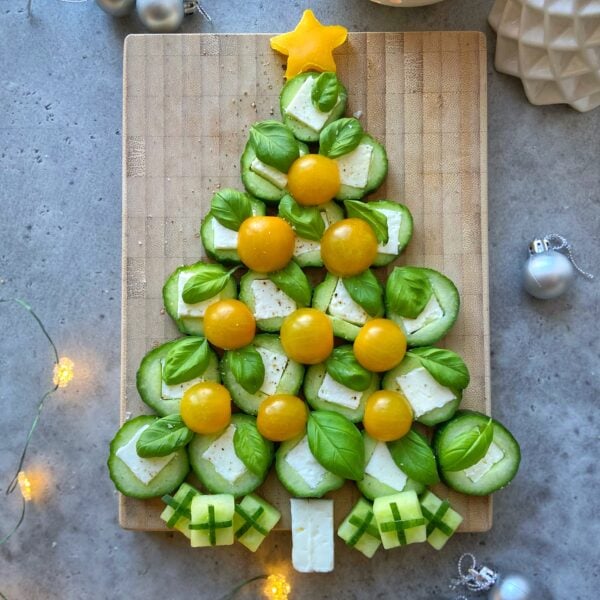 A festive Christmas tree shape made from cucumber slices, cheese, basil leaves, and yellow cherry tomatoes on a wooden board, with cheese and cucumber bites as gift decorations.