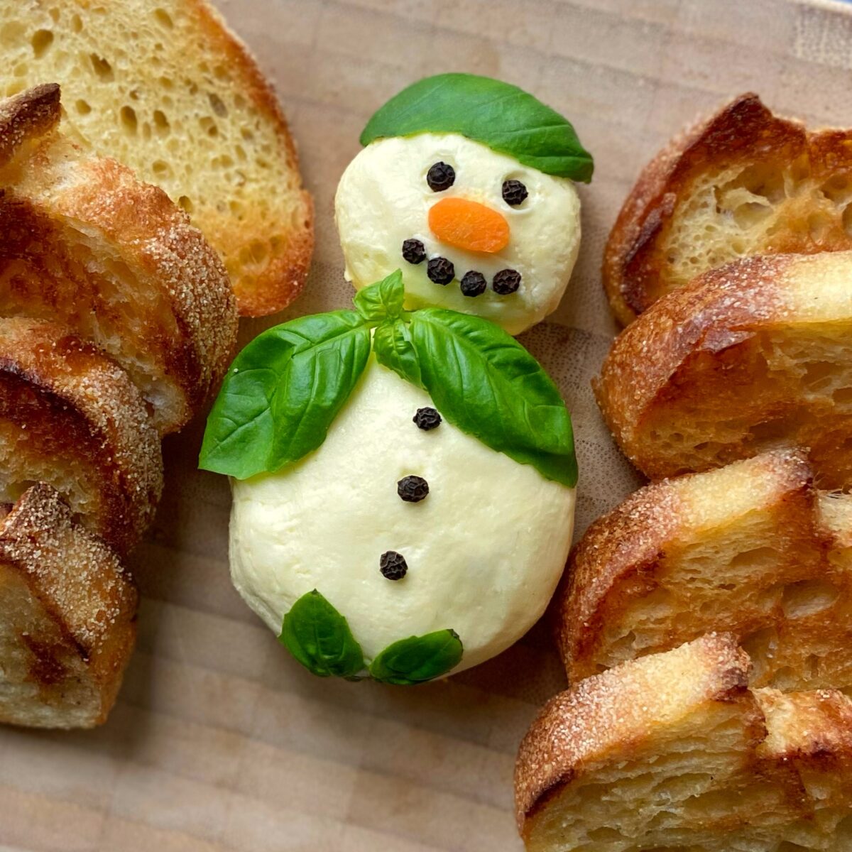 A closeup of a Whipped Butter Snowman, decorated with basil leaves, peppercorns, and a carrot nose, sits on a wooden surface surrounded by slices of toasted bread.