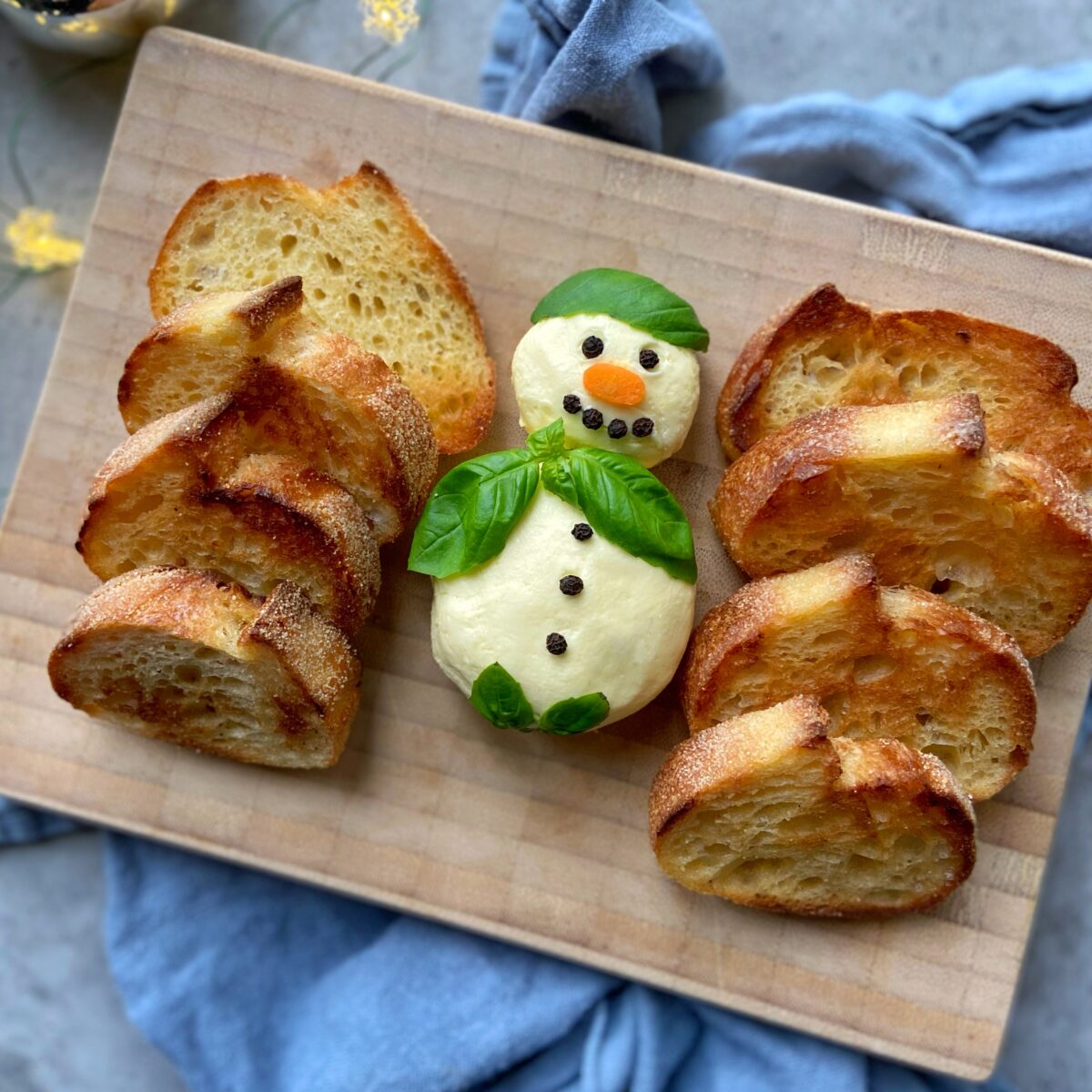 A snowman shaped out of butter decorated with basil, carrot, and peppercorns is on a wooden board, with slices of toasted bread around it.